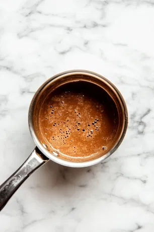 A small saucepan on a white marble cooktop with heavy cream, butter, and brown sugar melting over medium heat, then thickened with salt and vanilla.