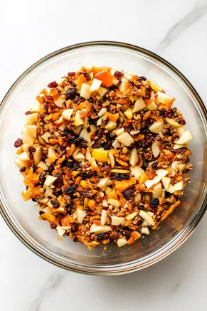 A large mixing bowl on the white marble cooktop, filled with mixed dried fruits, finely chopped candied peel, diced apple, fresh orange zest and juice, lemon zest and juice, and a splash of brandy. The bowl is covered, letting the ingredients marinate for at least 2 hours or overnight, developing rich flavors.