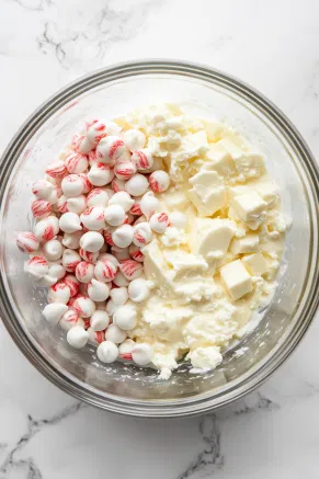 A large microwave-safe bowl filled with white chocolate chips, sweetened condensed milk, butter, and peppermint extract sits on a white marble cooktop. The mixture is partially melted and creamy, showing a smooth consistency as it is heated in short intervals to prevent overheating.