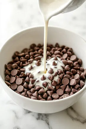 Chocolate chips in a heat-safe bowl on the white marble countertop, with hot cream being poured over them to create a smooth, glossy ganache.