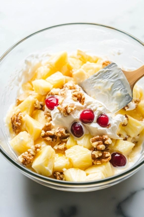 A large glass mixing bowl on the white marble cooktop is filled with crushed pineapple, lemon juice, thawed whipped topping, chopped cherries, sweetened condensed milk, and chopped walnuts. A spatula gently folds the vibrant, creamy mixture together.