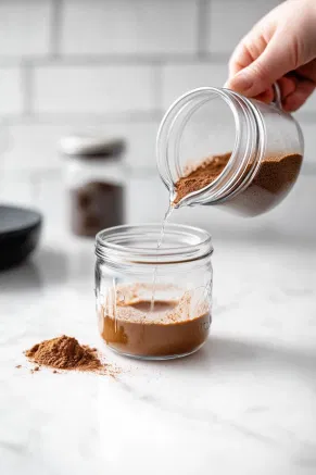 A sealable jar on a white marble cooktop being filled with warm water, instant coffee granules, and sugar, as a person closes the lid to shake the mixture.