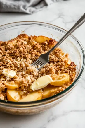 A large glass bowl on the white marble cooktop contains oats, flour, brown sugar, baking powder, and baking soda. Melted butter is being mixed in with a fork to create a crumbly topping.