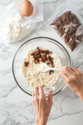 A medium mixing bowl on a white marble countertop with hands stirring together flour, brown sugar, and an egg, with bags of flour and brown sugar in the background.