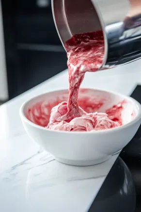 The strawberry mixture being poured into the freezer bowl of an ice cream maker on the white marble countertop.