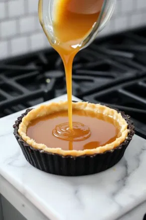 Golden caramel being poured evenly into the tart base on the white marble countertop, forming a smooth layer.