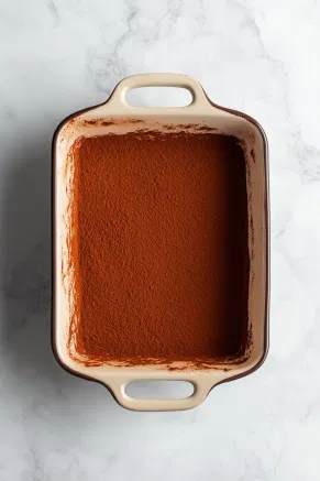 The bottom of a baking dish on the white marble cooktop, evenly dusted with a thin layer of cocoa powder, providing the base layer for the tiramisu.