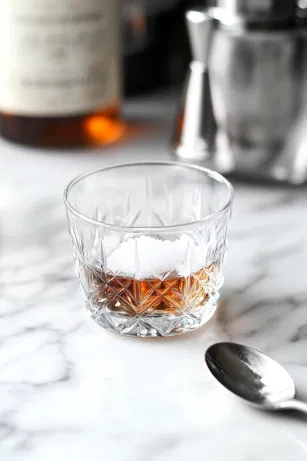 A clear glass mixing glass is positioned on the white marble cooktop, containing sugar and dashes of Angostura bitters at the bottom. A spoon is gently stirring as a small amount of water is added, dissolving the sugar until it becomes nearly clear, showing the careful preparation.