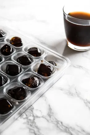 A tray of ice cube molds on a white marble countertop filled with strong black coffee, set to freeze until solid.