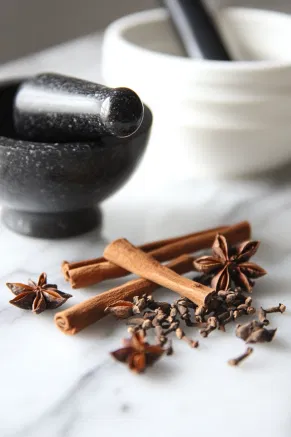 A pestle and mortar on a white marble cooktop, with star anise, nutmeg, cinnamon sticks, and whole cloves being roughly crushed, releasing warm, winter aromas. The crushed spices are being transferred into a jar with gin, ready for a 5-day infusion.