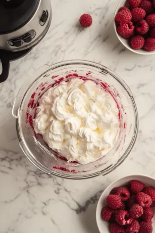 A food processor on the white marble cooktop shows raspberries and icing sugar being blended into a smooth sauce, which is then strained to remove seeds. In a separate bowl, double cream is whipped with icing sugar, a splash of sherry, and vanilla extract to soft peaks, creating a light and fluffy topping.