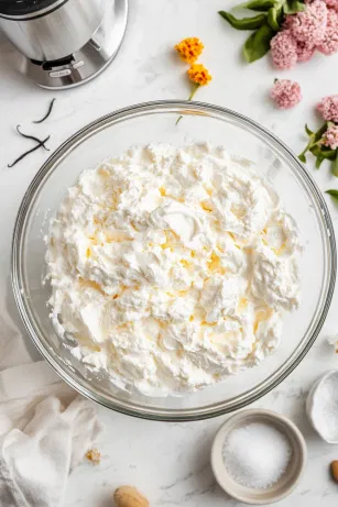 In a large mixing bowl on the white marble cooktop, cold double cream, icing sugar, and vanilla extract are whipped with an electric mixer on low to medium speed until soft peaks form. Once ready, the whipped cream is placed in the fridge to chill.