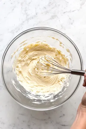 A large glass mixing bowl on the white marble cooktop with Angel Food cake mix being whisked into a smooth batter, ready for baking.