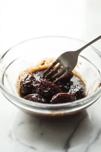 A small bowl on a white marble cooktop with boiling water poured over pitted Medjool dates, soaking for softening before mashing.