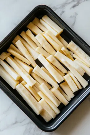 Peeled and chopped parsnips are arranged in a black roasting tin on a white marble cooktop. The pieces are finger-sized and uniform, ready for seasoning.