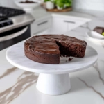 The completed gluten-free chocolate fudge cake on a cake stand on the white marble countertop, beautifully sliced and ready to be served.