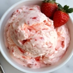 Scoops of creamy homemade strawberry ice cream served in a bowl on the white marble countertop, garnished with a fresh strawberry for a delightful presentation.