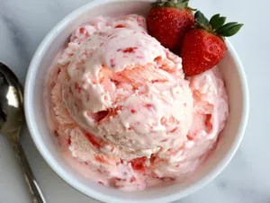 Scoops of creamy homemade strawberry ice cream served in a bowl on the white marble countertop, garnished with a fresh strawberry for a delightful presentation.