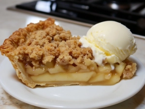 A warm slice of apple crumb pie is served on a dessert plate, placed on the white marble cooktop. A scoop of vanilla ice cream melts over the pie, creating an enticing dessert presentation.