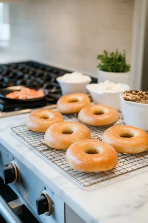 Freshly baked bagels cool on a wire rack on the white marble cooktop, ready for slicing and serving. Cream cheese, smoked salmon, and other toppings sit nearby, inviting a delicious spread.