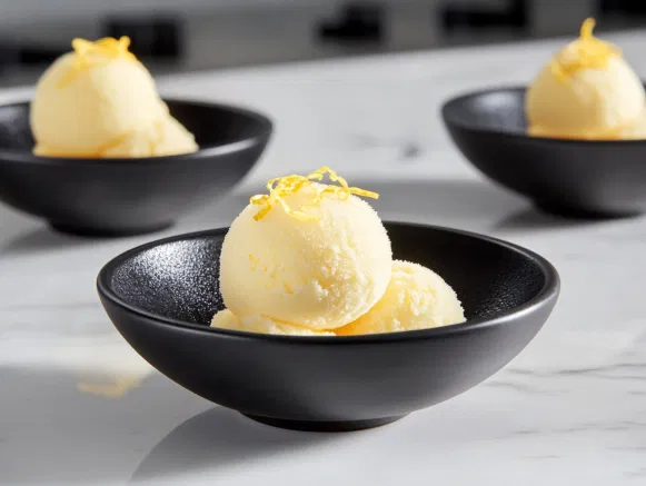 Scoops of lemon sorbet in black ceramic bowls on a white marble cooktop, garnished with fresh lemon zest for a colorful, refreshing presentation.