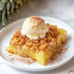 A small white plate with a serving of pineapple crisp, topped with a scoop of vanilla ice cream, placed on a white marble counter with a light sprinkle of cinnamon nearby.