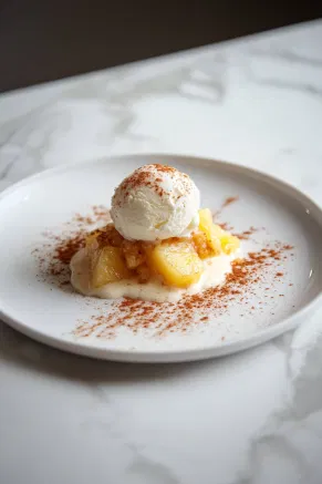 A small white plate with a serving of pineapple crisp, topped with a scoop of vanilla ice cream, placed on a white marble counter with a light sprinkle of cinnamon nearby.