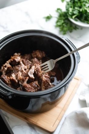 The black slow cooker on the white marble cooktop is shown with neck bones being transferred onto a wooden cutting board. A fork shreds the tender meat, while the empty slow cooker awaits the next ingredients.