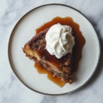 A slice of sticky toffee pudding served on a plate on the white marble countertop, topped with a generous drizzle of toffee sauce and a dollop of vegan whipped cream.