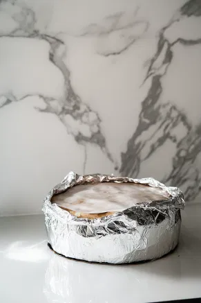 The cake is wrapped in parchment paper and foil, ready for storage in an airtight container on the white marble cooktop. It can be ‘fed’ with additional rum or brandy every 1-2 weeks for enhanced flavor.