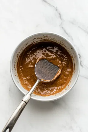 The prepared baking pan sits on the white marble cooktop as the batter is poured through a sieve into the pan, ensuring a smooth texture by removing any lumps.