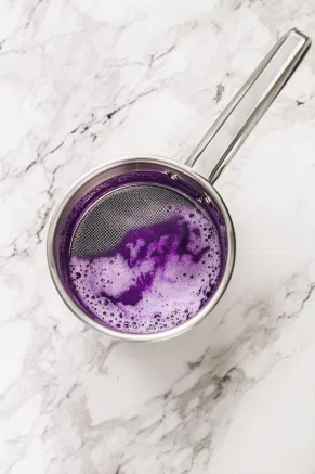 The vibrant purple liquid being strained through a sieve into a pitcher on the white marble countertop, separating it from the solids.