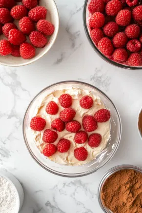 A final layer of the cream cheese mixture is added to the trifle bowl on the white marble cooktop. The remaining whipped cream is spread over the top, and a sifter dusts cocoa powder over the surface, giving a polished and inviting finish.