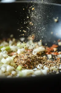 Salt, poultry seasoning, and black pepper are added to the skillet with the sautéed onion and celery. The mixture cooks for 1 more minute, becoming fragrant on the white marble cooktop