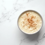 A serving bowl of warm rice pudding sits on the white marble cooktop, sprinkled with ground cinnamon for garnish. The dish is ready to be enjoyed, served warm or cold.