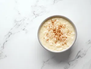 A serving bowl of warm rice pudding sits on the white marble cooktop, sprinkled with ground cinnamon for garnish. The dish is ready to be enjoyed, served warm or cold.