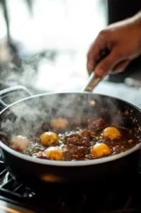 The browned meat is returned to the Dutch oven on the white marble cooktop, with the sauce and remaining broth poured over it. The mixture is brought to a simmer, then covered and placed in a preheated 300°F oven to braise for 3 hours until tender.