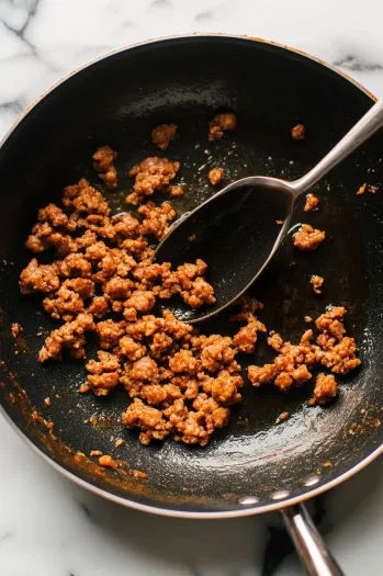 A large skillet on the white marble cooktop as breakfast sausage cooks over medium-high heat, browning and crumbling. A slotted spoon is ready to transfer the cooked sausage to a large mixing bowl.