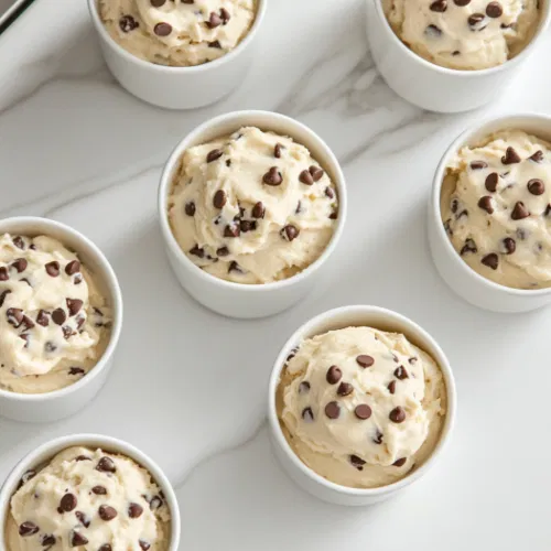 Spoonfuls of edible cookie dough served in small bowls on the white marble cooktop, with chocolate chips visible throughout the dough, ready to be enjoyed directly from the bowl.