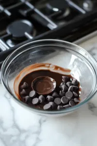 A microwave-safe bowl on the white marble cooktop with dark chocolate chips and 1 tablespoon of coconut oil. The chocolate is being melted in the microwave in 20-second increments, stirred to a smooth consistency.