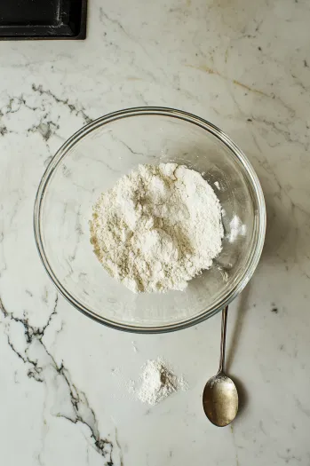 A large glass mixing bowl on the white marble cooktop with flour, sugar, baking powder, and salt being whisked together. A spoon is positioned nearby, ready to combine the ingredients