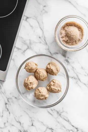 A hand rolling 1.5-tablespoon-sized balls of snickerdoodle cookie dough and coating them in cinnamon-sugar topping before placing them on the baking sheet.