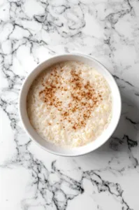 A serving bowl of warm rice pudding sits on the white marble cooktop, sprinkled with ground cinnamon for garnish. The dish is ready to be enjoyed, served warm or cold.