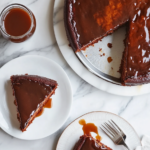 The Swedish Apple Cake cools on the white marble cooktop, its springform pan removed to reveal a caramel-cracked top. Slices are plated with forks, accompanied by a small pitcher of extra caramel sauce for drizzling.