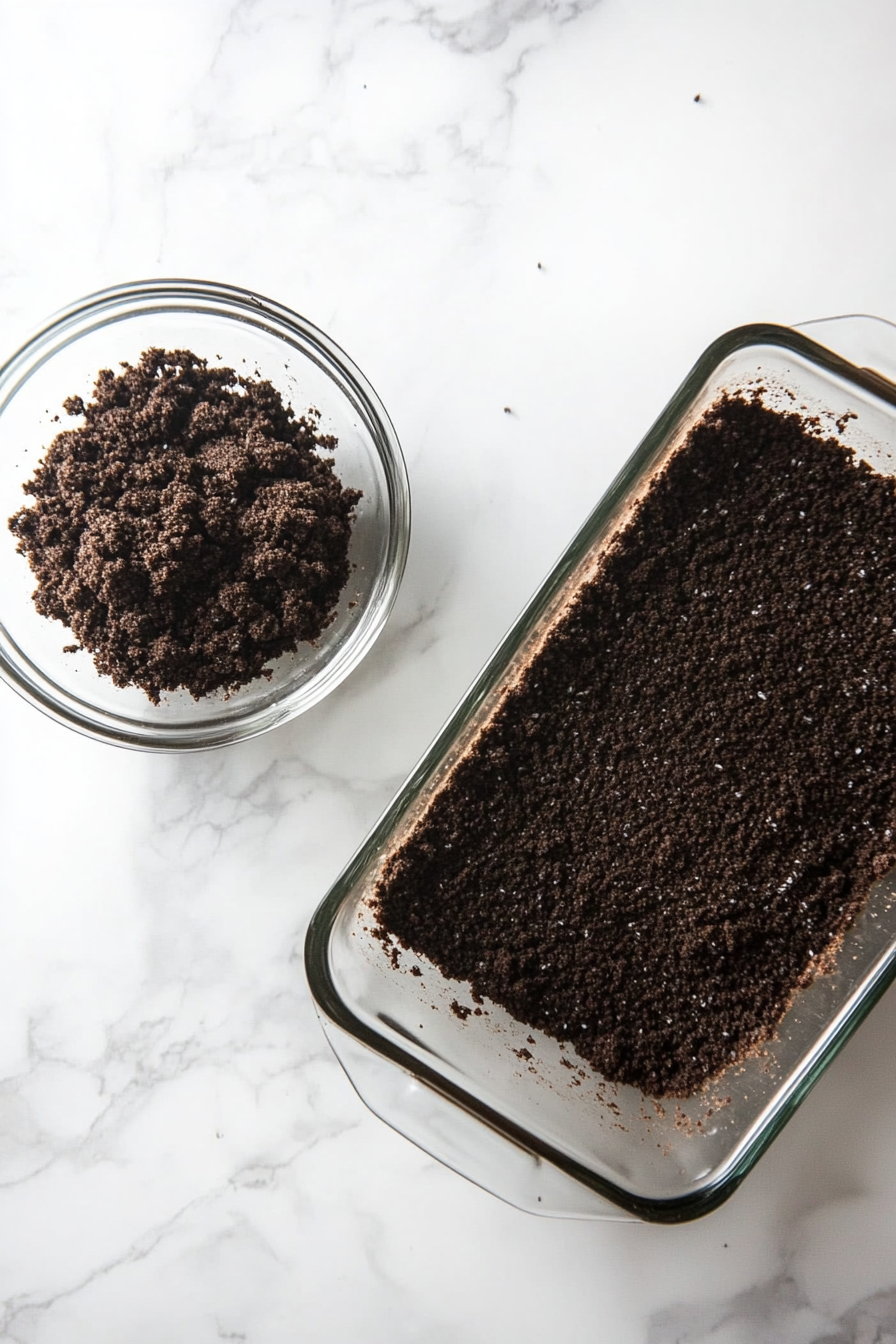 A medium glass bowl on a white marble countertop contains crushed Oreo crumbs mixed with espresso powder and melted butter. The mixture appears dark and moist with a uniform texture. Next to the bowl, the loaf pan holds the pressed crust, forming a smooth and compact layer of dark crumbs.