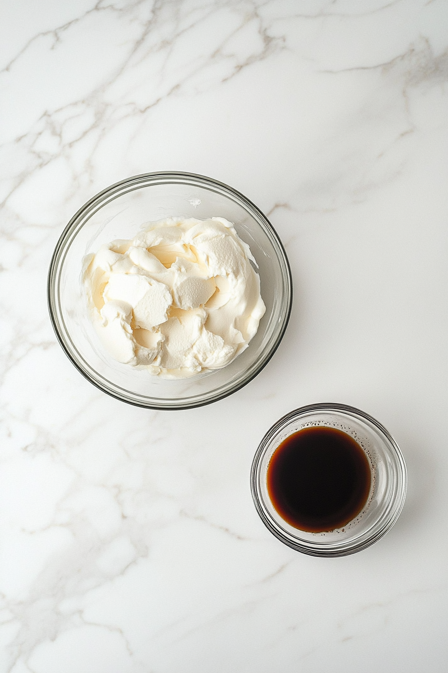 A medium glass bowl on the white marble countertop contains cream cheese and sugar beaten into a fluffy white mixture. A small bowl nearby holds espresso powder dissolved in warm water, ready to be added. The setup remains minimal and clean.