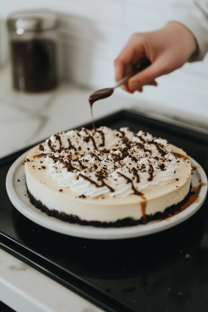 The cheesecake sits on the white marble cooktop, with freshly whipped cream elegantly piped onto the top. Crushed Oreos are sprinkled over the whipped cream, and a drizzle of hot fudge sauce is added as a decorative garnish. The finished cheesecake looks decadent and ready to be served.