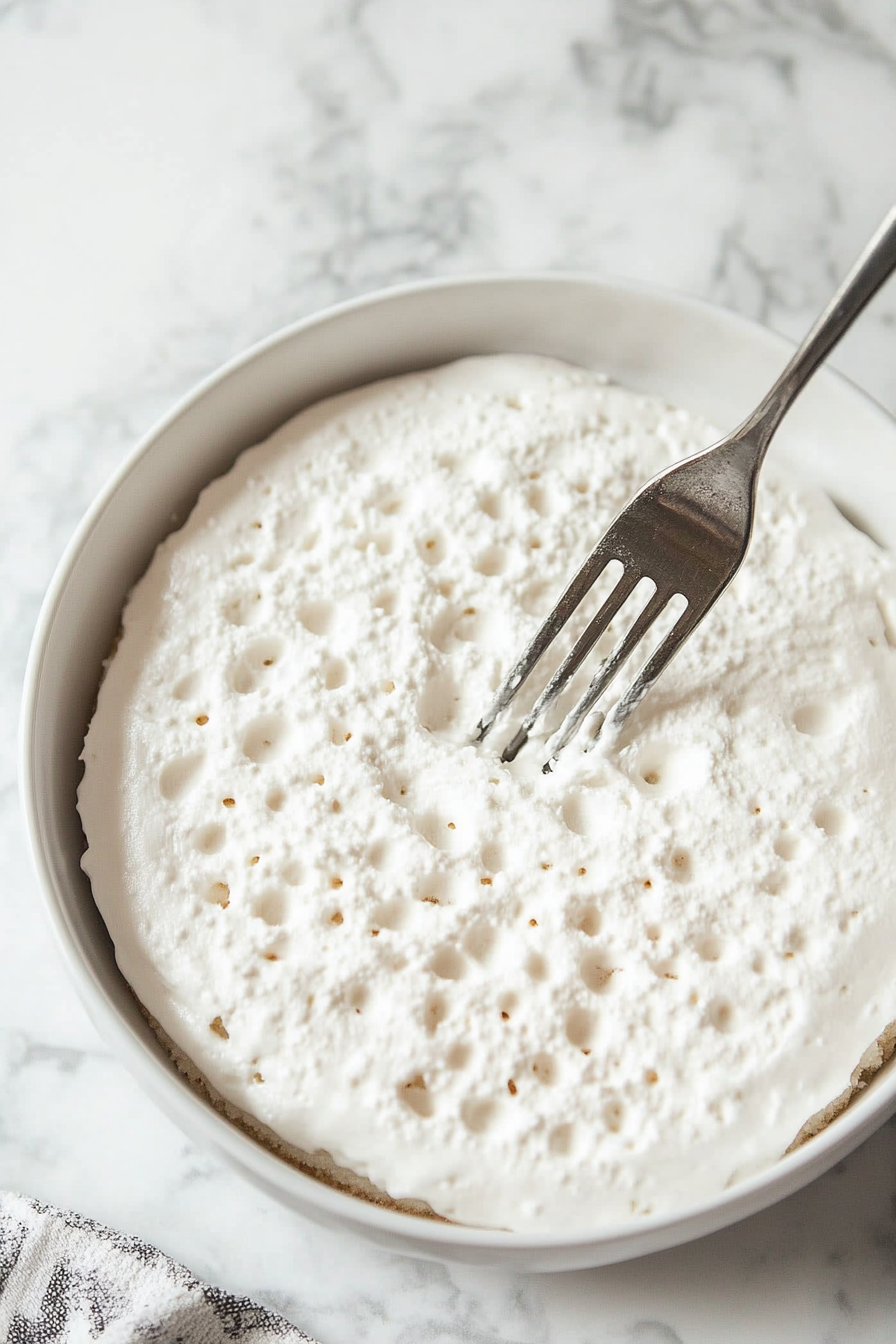 A cooled white cake on a white marble cooktop, with a fork creating evenly spaced holes across its surface.