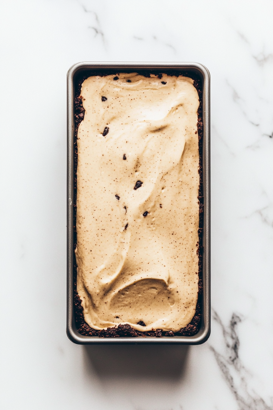 The loaf pan with the Oreo crust is now filled with the light beige coffee cheesecake mixture. The surface of the filling is smooth and leveled, with no bubbles. The pan is placed on the white marble countertop.