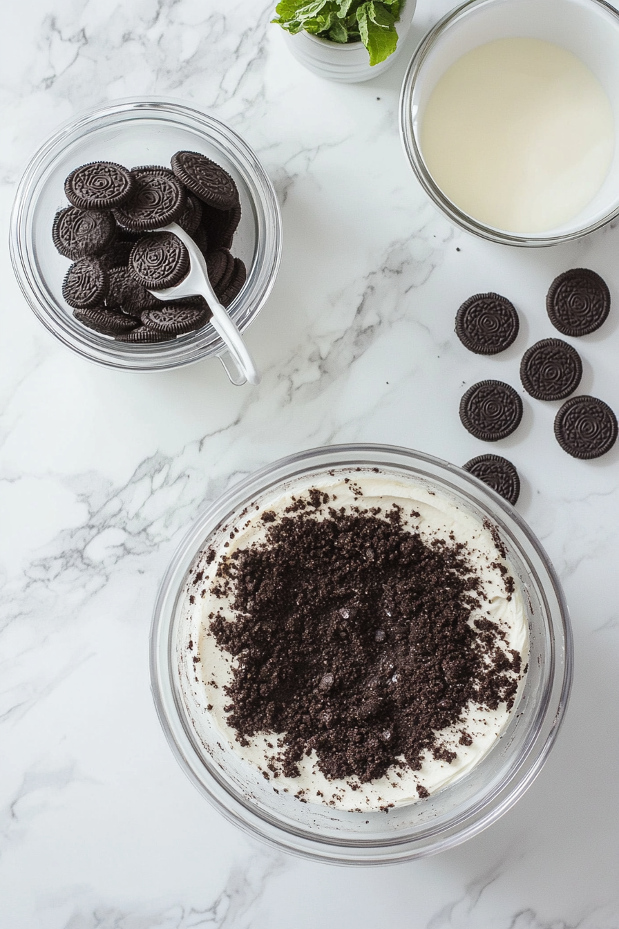 A 9-inch springform pan with the bottom lined with parchment paper sits on a white marble cooktop. Oreos are being crushed into fine crumbs in a food processor, and melted butter is ready to mix. The crushed Oreos are combined with butter in a bowl, and the mixture is pressed into the bottom and sides of the pan.